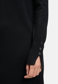 Maglione knit nero con polsini a coste, caratterizzato da tre bottoni decorativi neri sulla manica e una leggera apertura per un tocco in più.