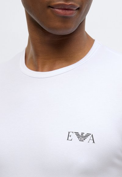 Bílé bavlněné tričko s kulatým výstřihem, s malým šedým logem na hrudi. Hladká textura, přiléhavý střih.