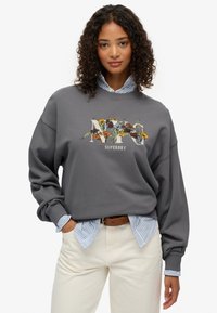 Sudadera gris con gráfico floral "NY", cuello redondo, hombros caídos, encima de una camisa a rayas azul, combinada con pantalones crema.