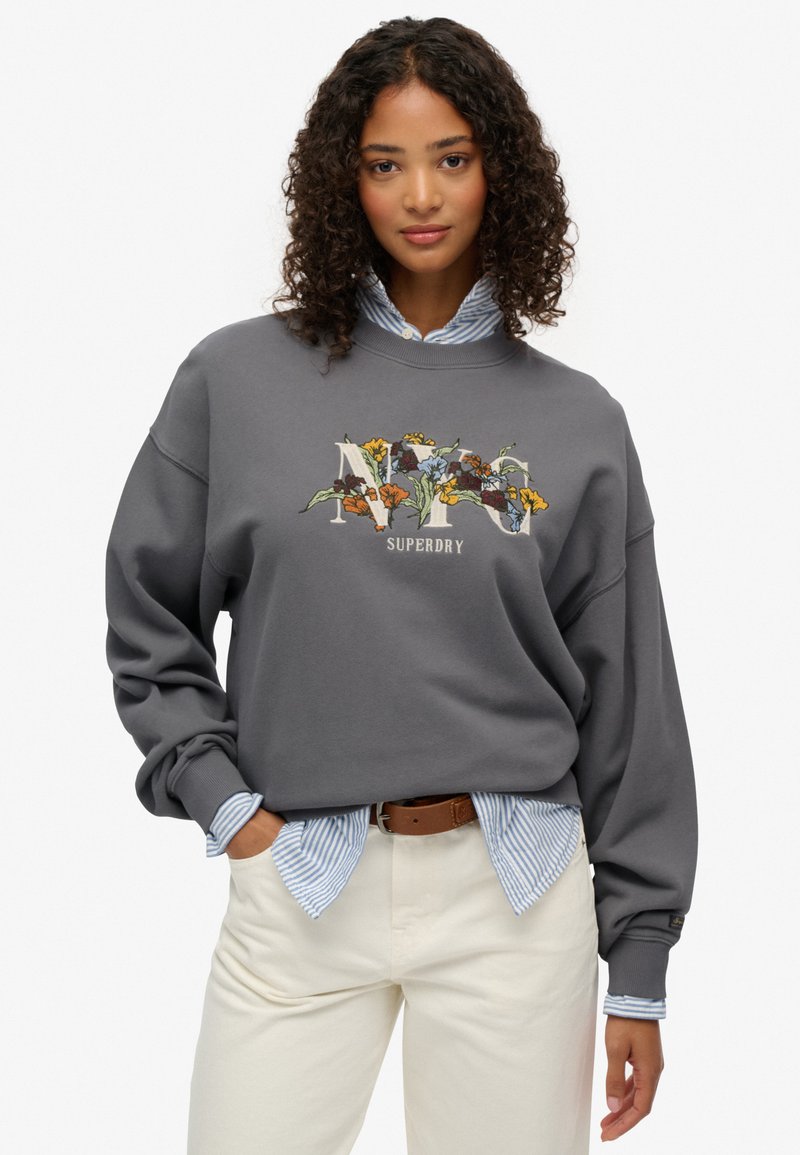 Sudadera gris con gráfico floral "NY", cuello redondo, hombros caídos, encima de una camisa a rayas azul, combinada con pantalones crema.
