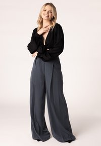 Zwarte blouse met lange mouwen, een diepe V-hals en pofmouwen. Grijze, hoge wijde broek met plooien en een vloeiende textuur.