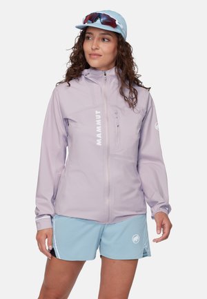 Kvinde iført en lyselilla lynlåstrøje, lyseblå shorts, en lyseblå kasket og solbriller placeret på kasketten.