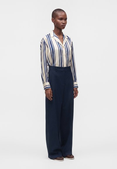 Femme debout portant une chemise boutonnée rayée verticale crème et bleue, rentrée dans un pantalon large taille haute bleu marine et des chaussures marron.
