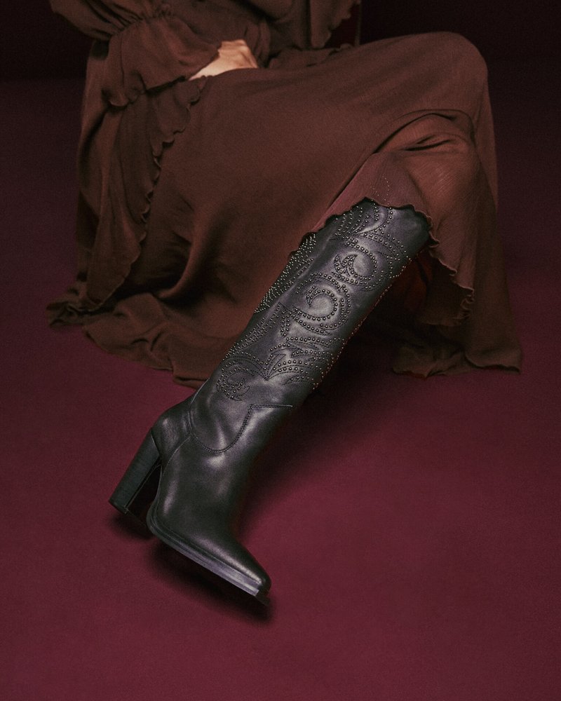 Bota de cuero negro hasta la rodilla con tacón cuadrado, que presenta patrones en relieve de espirales y acentos de tachuelas, colocada sobre un fondo en color burdeos.