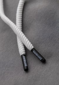 Szara tkanina w tle z białymi plecionymi sznurówkami, wyposażonymi w czarne plastikowe agle z wytłoczonym napisem "OMBRÉ", co ułatwia ich sznurowanie.