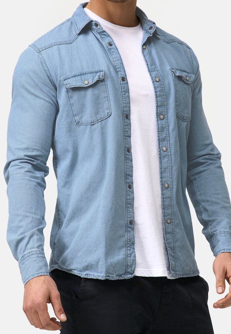 Homme portant une chemise en jean bleu clair boutonnée sur un t-shirt blanc uni, associée à un pantalon noir, debout sur un fond blanc.