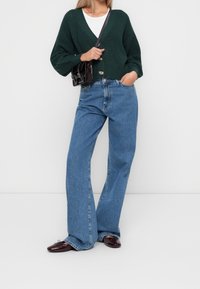 Zielony sweter z dzianiny z dekoltem w serek i dwoma guzikami, zestawiony z niebieskimi spodniami z wysokim stanem i szerokimi nogawkami oraz ciemnymi mokasynami. Mała czarna torebka.