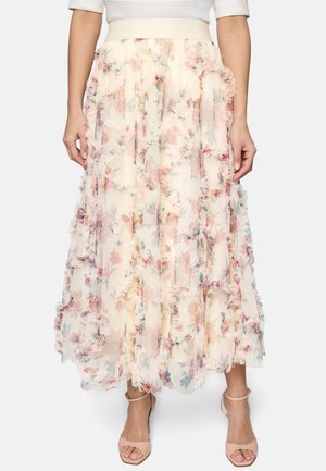 Femme portant une jupe longue crème à motifs floraux avec des couches volantées et des talons beiges à bride autour de la cheville, assortie à un haut blanc.