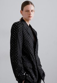 Black blazer featuring a textured surface with circular metallic accents, sharp lapels, and a tailored fit.