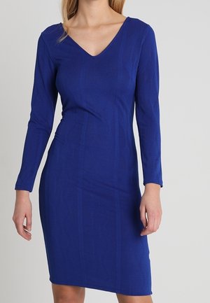 Femme portant une robe bleue ajustée, à manches longues, longueur genou, avec un col en V, sur un fond clair uni.