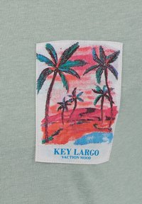 Naszywka z palmami w stylu akwarelowym, czerwonym plażą, niebieską wodą oraz napisem "KLIMAT WAKACJI KEY LARGO" na jasnooliwkowym tle.