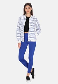 Jachetă bomber albă cu fermoar, cu modele albastre, top croppat negru și colanți albaștri. Prezintă un design ajustat și manșete elastice. Adidași negri.