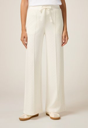 Pantalons larges crème avec un motif texturé à pois. Comprend une taille à cordon de serrage et des poches latérales. Portés avec des chaussures beige à enfiler.