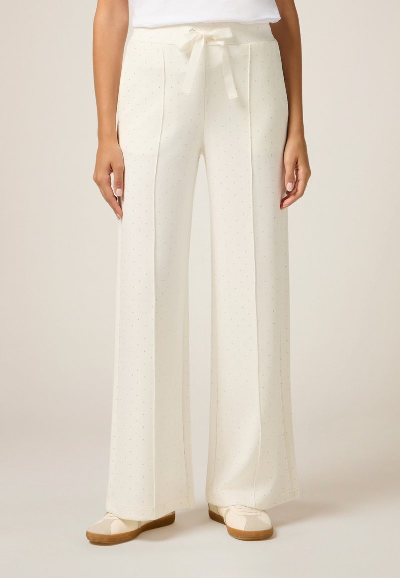 Pantalons larges crème avec un motif texturé à pois. Comprend une taille à cordon de serrage et des poches latérales. Portés avec des chaussures beige à enfiler.