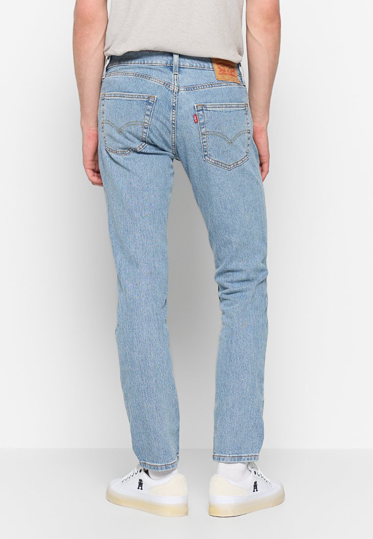Persona vistiendo jeans rectos de color azul claro, calcetines blancos y zapatillas blancas con suelas beige, de pie frente a una pared blanca.