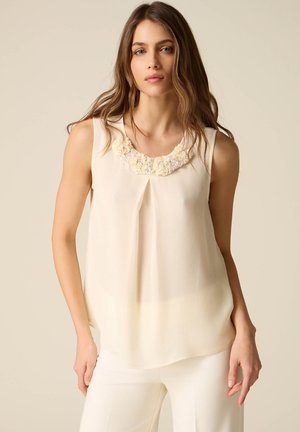 Žena v krémové bezrukávové blůze s květinovou výšivkou na výstřihu a krémové kalhoty, stojící před neutrálním pozadím.