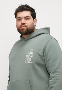 Zeleni hoodie od pamuka s grafikom i natpisom "Opusti se, ništa se neće dogoditi" u bijeloj boji. Kapica s kengurufrontom.