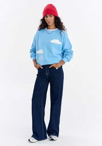 Jasnoniebieska bluza z białymi motywami chmur, zestawiona z ciemnoniebieskimi spodniami o szerokich nogawkach i białymi tenisówkami. Na głowie czerwona dziergana czapka.