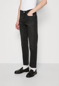 Jeans in denim nero con una vestibilità a gamba dritta, dotati di cinque tasche, chiusura con zip e una texture leggermente sbiadita. Abbinati a mocassini neri e calzini bianchi.