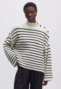 Gestreifeter Pullover in Weiß und Schwarz, mit hohem Kragen, lockerer Passform und Knopfdetails an der Schulter. Strukturierter Strickstoff.