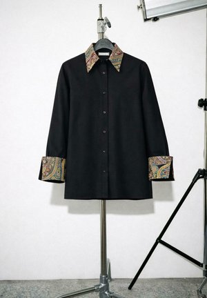 Sort langærmet skjorte med knapper foran, med farverigt paisley-mønster på krave og manchetter, vist på et metalstativ i et studie.