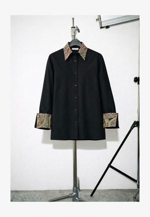 Sort langærmet skjorte med knapper foran, med farverigt paisley-mønster på krave og manchetter, vist på et metalstativ i et studie.