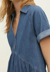 Denim jurk met een geknoopte halslijn, knoopsluiting aan de voorkant, korte opgerolde mouwen en een geplooide rok. De stof is lichtblauw met een gladde textuur.