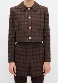 Brun ternet cropped jakke med krave, med guldknapper og en tekstureret finish. Inkluderer to forlommer og lange ærmer.