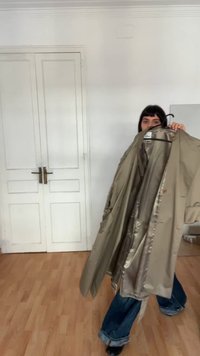 Beżowy trencz o gładkiej, błyszczącej fakturze i luźnym kroju. Posiada kołnierz, szerokie rękawy i duże guziki.