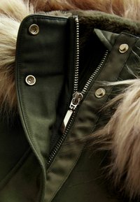 Olivgrüner Parka mit silbernem Reißverschluss, Metallschnallen und einem Kunstfellbesatz. Strukturierter Stoff mit horizontalen Rippendetails in der Nähe des Reißverschlusses.