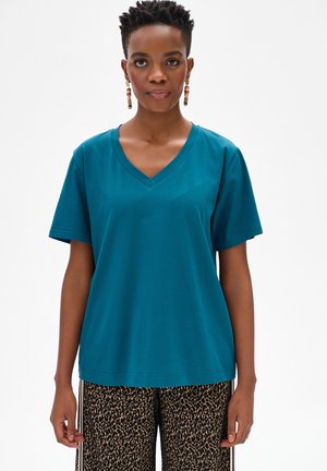 Kobieta ubrana w zielononiebieską koszulkę z krótkim rękawem i dekoltem w serek oraz spodnie w panterkę, stojąca na tle jednolitego białego tła.