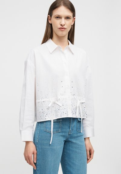 Femme portant une chemise blanche à boutons avec de petites taches noires et bleues, nouée à la taille, associée à un jean en denim bleu.