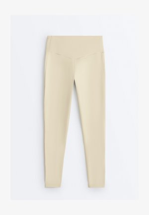 Bež legice z visokim pasom in V-oblika šivom na sprednji strani, položene razgrnjene na belem ozadju.