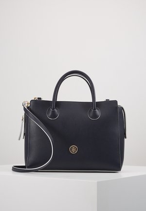 Sort læderhåndtaske med dobbelthanke, aftagelig skulderrem og lille gyldent rundt logo foran, placeret på hvid overflade.