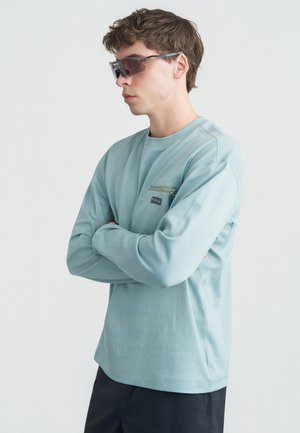 Noor mees, kes kannab helesinist pika varrukaga särki, millel on lukuga rinnatasku, ja tumedaid päikeseprille, seisab valge tausta ees käed kokku pandud.