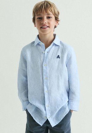 Niño rubio está de pie con las manos en los bolsillos, vistiendo una camisa azul claro abotonada con un pequeño emblema oscuro en el pecho.