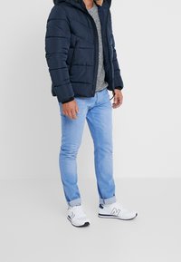 Donkerblauwe pufferjas met capuchon, grijze gestructureerde trui, lichtblauwe spijkerbroek en witte sneakers met blauwe accenten. Voorzien van ritssluiting en horizontale stiknaden.