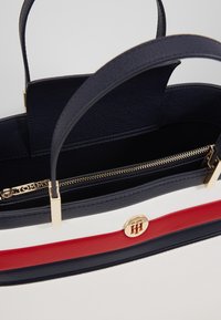 Laadukkaasta tummansinisestä nahasta valmistettu käsilaukku, jossa on valkoinen ja punainen raita, ylhäältä suljettava vetoketju, kultaiset yksityiskohdat ja kaksoiskahvat, sekä teksturoitu pinta.