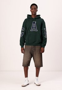 Grønn hettegenser med store hvite bokstaver "A" og "G" på ermene, iført sammen med løse, slitte brune shorts og hvite sneakers.