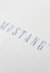 Witte katoenen t-shirt met grote blauwe bloemenpatroon "MUSTANG" tekst op de voorkant. Gladde textuur, relaxte fit, minimalistisch ontwerp.