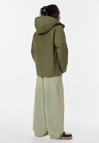 Oliven grøn, hættebeklædt pufferjakke; letvægtigt, let glansende stof. Sammen med løse, falmede beige denimbukser og brune sko.