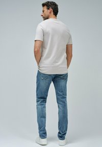 Hombre de pie de espaldas, vistiendo una camisa de manga corta beige claro, jeans azules y zapatillas blancas, frente a un fondo gris liso.