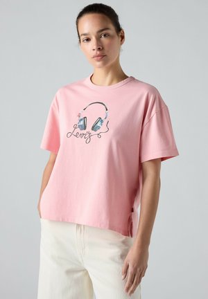 Vrouw draagt een losse roze t-shirt met geïllustreerde koptelefoons en cursieve tekst, gecombineerd met ecru broek, staand tegen een effen achtergrond.