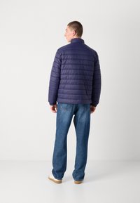 Marineblå pufferjakke med horisontal søm, høy krage og elastiske mansjetter, sammen med løstsittende denimjeans og hvite joggesko.