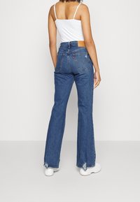 Femme portant un jean droit taille haute bleu avec des déchirures légères au niveau des ourlets, un débardeur blanc et des baskets compensées blanches, se tenant contre un mur blanc.