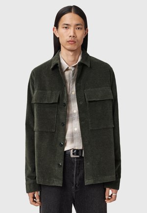 Dark green corduroy jacket with a button-up front, two chest pockets, and a relaxed fit. Worn over a light plaid shirt.