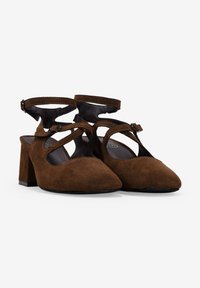 Zapatos altos de ante marrón con punta afilada, laterales abiertos y tirantes para los tobillos. Los tacones cuadrados ofrecen estabilidad. Textura interior suave.