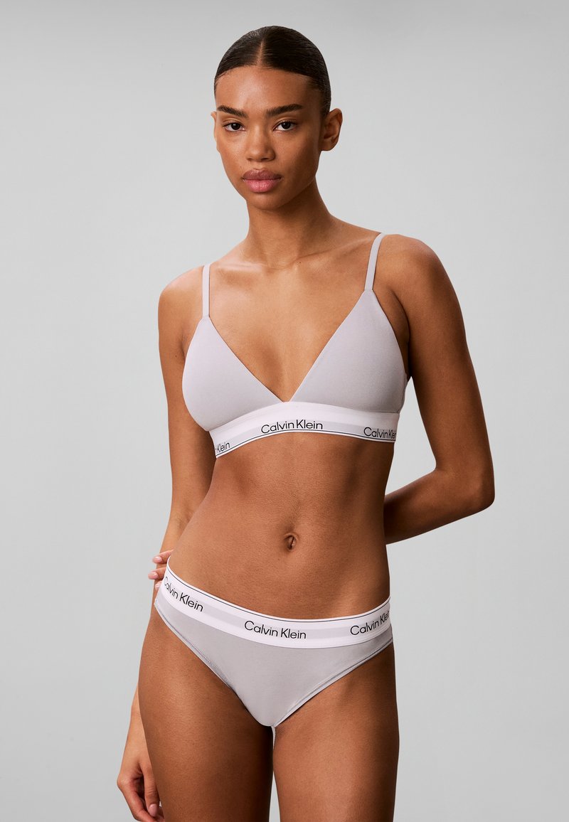 Γυναίκα που φοράει ανοιχτό γκρι bralette Calvin Klein και ταιριαστό εσώρουχο με ελαστικές ταινίες με λογότυπο, στέκεται μπροστά σε απλό γκρι φόντο.