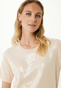 Pull court à manches courtes couleur beige clair avec un motif texturé en sequins. Encolure ronde et tissu doux. Design simple avec une légère brillance.