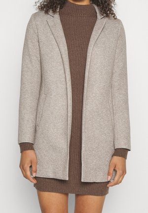 Femme portant un blazer beige sur une robe en maille marron côtelée à manches longues, debout devant un fond blanc uni.